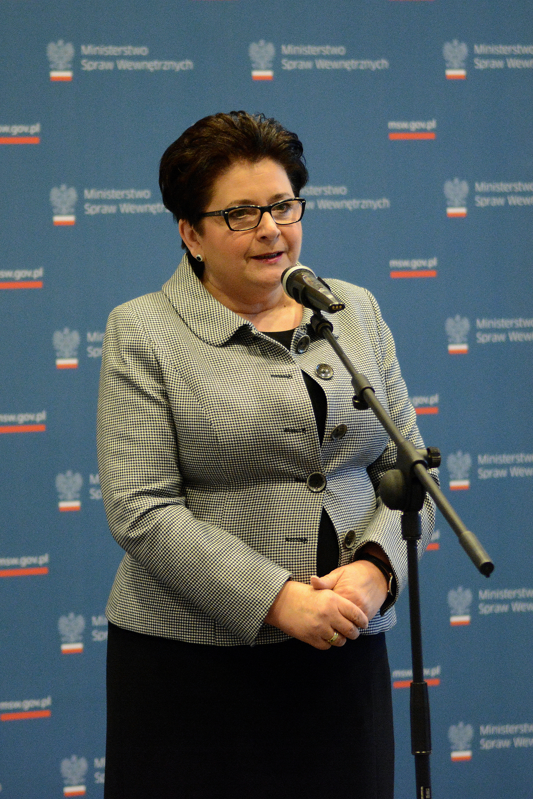 Zdjęcie kobiety Teresy Piotrowskiej. Kobieta w wieku około 55 lat. Stoi przed mikrofonem. Włosy czarne krótkie, zaczesane do tyłu. Czoło wysokie, gładkie. Okulary w czarnych grubych oprawkach. Nos mały, usta wąskie. Twarz okrągła. Uszy małe, przylegające. Dłonie splecione, wsparte na podbrzuszu. Ubrana w szarą marynarkę oraz czarną sukienkę.
