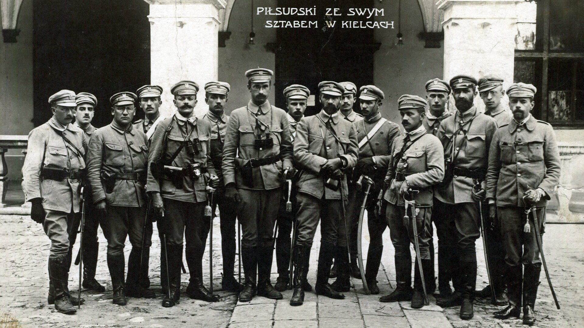 Czarno-biała fotografia przedstawia grupę 16 żołnierzy ubranych w mundury polskich legionistów. Każdy z nich ma przy pasie szablę. W centrum stoi Józef Piłsudski, po jego prawej i lewej stronie stoją członkowie sztabu. U góry zdjęcia widnieje napis: „PIŁSUDSKI ZE SWYM SZTABEM W KIELCACH”. Zdjęcie zrobiono przed pałacem. Grupa stoi ustawiona na tle pałacowego wejścia ozdobionego dwoma kolumnami. Pod zdjęciem zamieszczono informację o okolicznościach, w jakich zostało ono zrobione i przedstawionych na nim postaciach: „ Sztab oficerów legionowych w Kielcach, rok 1914. Od lewej: Ignacy Boerner, Zygmunt Sulistrowski, Aleksander Litwinowicz, Władysław Stryjeński, Michał Sokolnicki, NN, Kazimierz Sosnkowski, Michał Fuksiewicz, Józef Piłsudski, Michał Sawicki, Władysław Belina-Prażmowski, Mieczysław Trojanowski, Julian Stachiewicz, Walery Sławek, Roman Horoszkiewicz i Gustaw Daniłowski.”