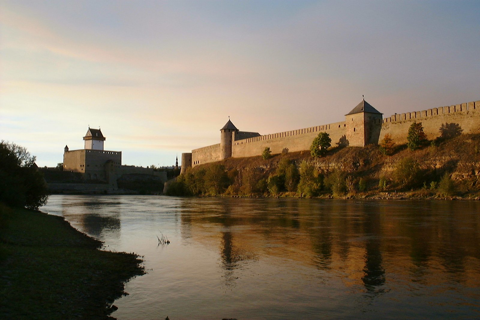 Zdjęcie robione z przeciwnego brzegu rzeki. Na pionowym brzegu rzeki stoi kamienny, szary mur obronny, w którym są dwie baszty. Jedna prostokątna, jedna okrągła. W basztach są wąskie okna. Dalej, po przeciwnejj stronie rzeki, nad jej brzegiem stoi biała, kwadratowa wieża otoczona murem obronnym
