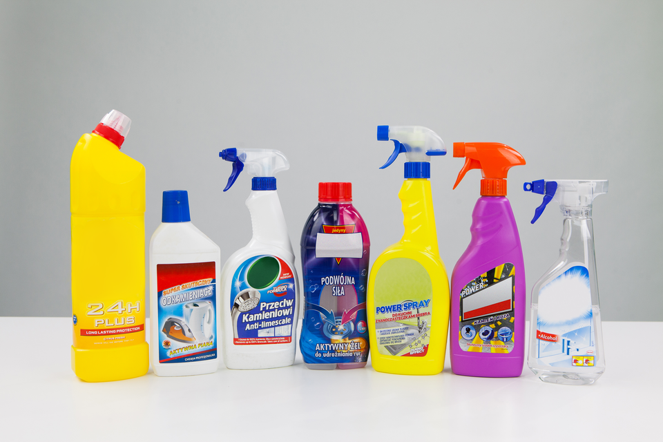 Zdjęcie przedstawia popularne środki czystości w różnych pojemnikach. Detergenty w postaci płynu są w pięciu plastikowych butelkach ułożonych w półokrąg. Po lewej stronie dwie butelki zakończone są ręcznymi spryskiwaczami. Trzecia butelka zakończona pionowo ustawionym dozownikiem, pozostałe dwie mają kolejno korek i zakrętkę z zabezpieczeniem przed otwarciem przez dzieci. Przed butelkami, na pierwszym planie pionowo stojące pudełko. Na prawo od pudełka plastikowy mniejszy okrągły pojemnik z zakrętką.