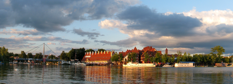 Fotografia prezentuje panoramę wybrzeża portowego w Mikołajkach na Mazurach. Na wybrzeżu widoczne zabudowania z czerwonymi dachami oraz cumujące przy brzegu statki i żaglówki.
