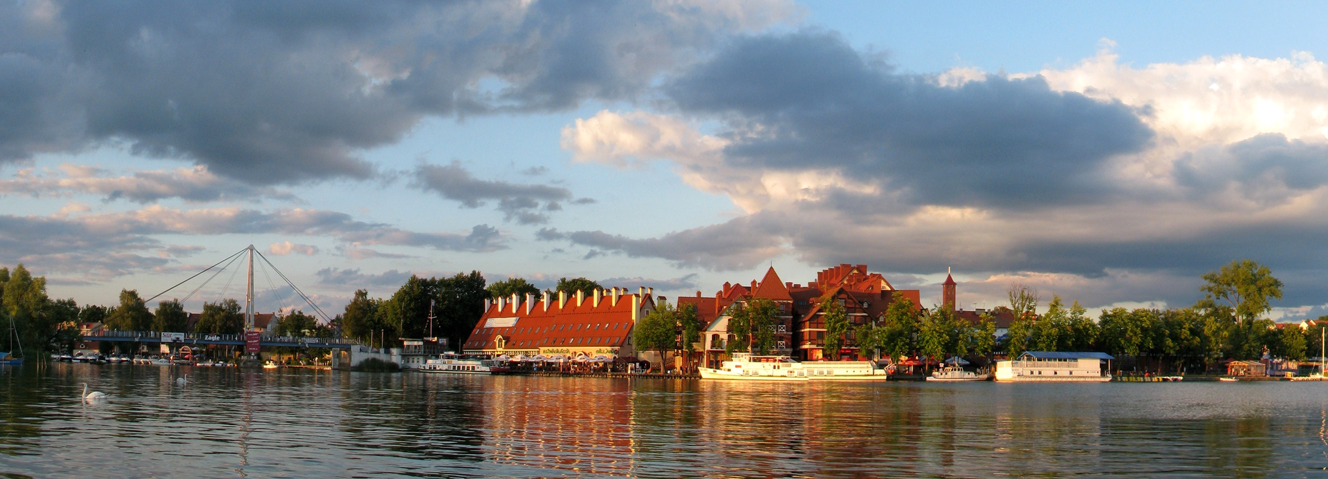 Fotografia prezentuje panoramę wybrzeża portowego w Mikołajkach na Mazurach. Na wybrzeżu widoczne zabudowania z czerwonymi dachami oraz cumujące przy brzegu statki i żaglówki.