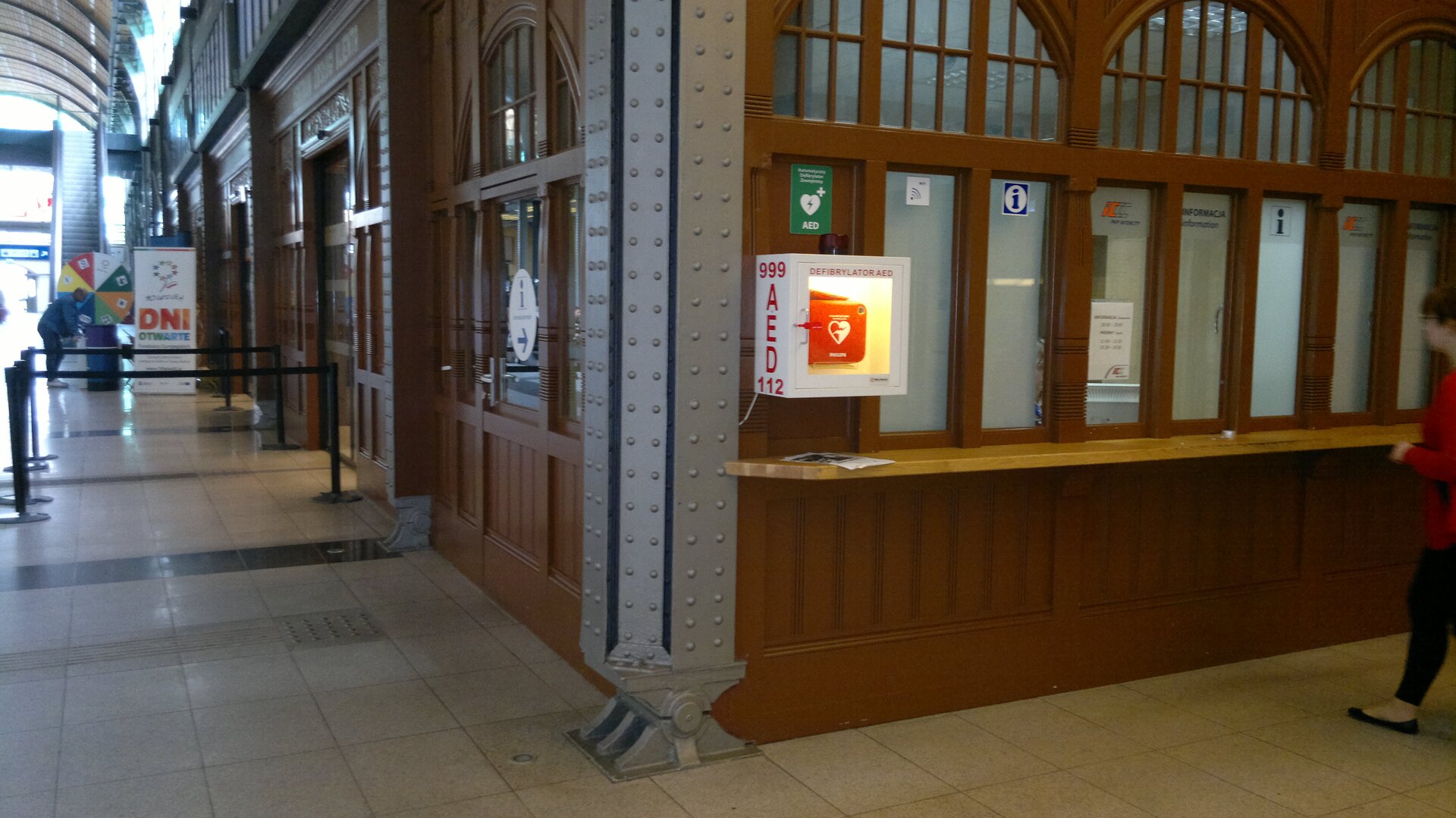 Kolorowe zdjęcie przedstawia fragment dworca kolejowego we Wrocławiu. Na pierwszym planie okienka z nalepkami informacja. Na lewo od okienek, na rogu ściany, mała biała oszklona szafka z defibrylatorem wewnątrz. Szafka w kształcie prostopadłościanu, oświetlona wewnątrz. Wymiary to 50 centymetrów wysokość i 20 centymetrów głębokość szafki. Na lewym boku szafki czerwony napis AED oraz numery telefonów. Na górze 999, na dole 112. Za rogiem ściany kasy biletowe. Kasy umieszczone jedna za drugą wzdłuż ściany budynku. Kasy znajdują się w głębi budynku. Wszystkie ściany wykonane są z ciemnobrązowego drewna. Okienka w kształcie pionowych prostokątów. Każde trzy okienka zakończone zwieńczającym półokrągłym oknem powyżej.