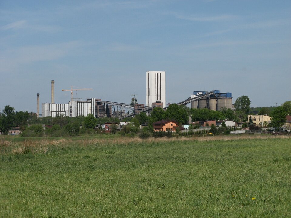 Zdjęcie przedstawia budynki kopalni węgla kamiennego Halemba w Rudzie Śląskiej. Słoneczny dzień, lato. Na pierwszym planie duża zielona polana. Na linii horyzontu, w planie średnim niskie budynki mieszkalne, a za nimi drzewa. Za drzewami budynki kopalni. W centrum zdjęcia, najwyższa budowla kopalni, biały budynek w kształcie pionowego prostopadłościanu. Jest to wieża wyciągowa. Na lewo dwa duże budynki, zakład przeróbczy. Ściany budynków pokryte pionowymi prostokątnymi białymi panelami. Za budynkami dwa wysokie okrągłe kominy wykonane z czerwonej cegły. Między kominami a wieżą wyciągową, znajduje się szyb górniczy wykonany z belek stalowych. Na szczycie szybu dwa koła wyciągowe. Po prawej stronie cztery wysokie budowle w kształcie walców o średnicy około dziesięciu metrów w szarym kolorze. To zbiorniki na węgiel. Wysokość budynków to połowa wysokości wieży wyciągowej. Dwa taśmociągi sięgają aż do dachu zbiorników na węgiel.