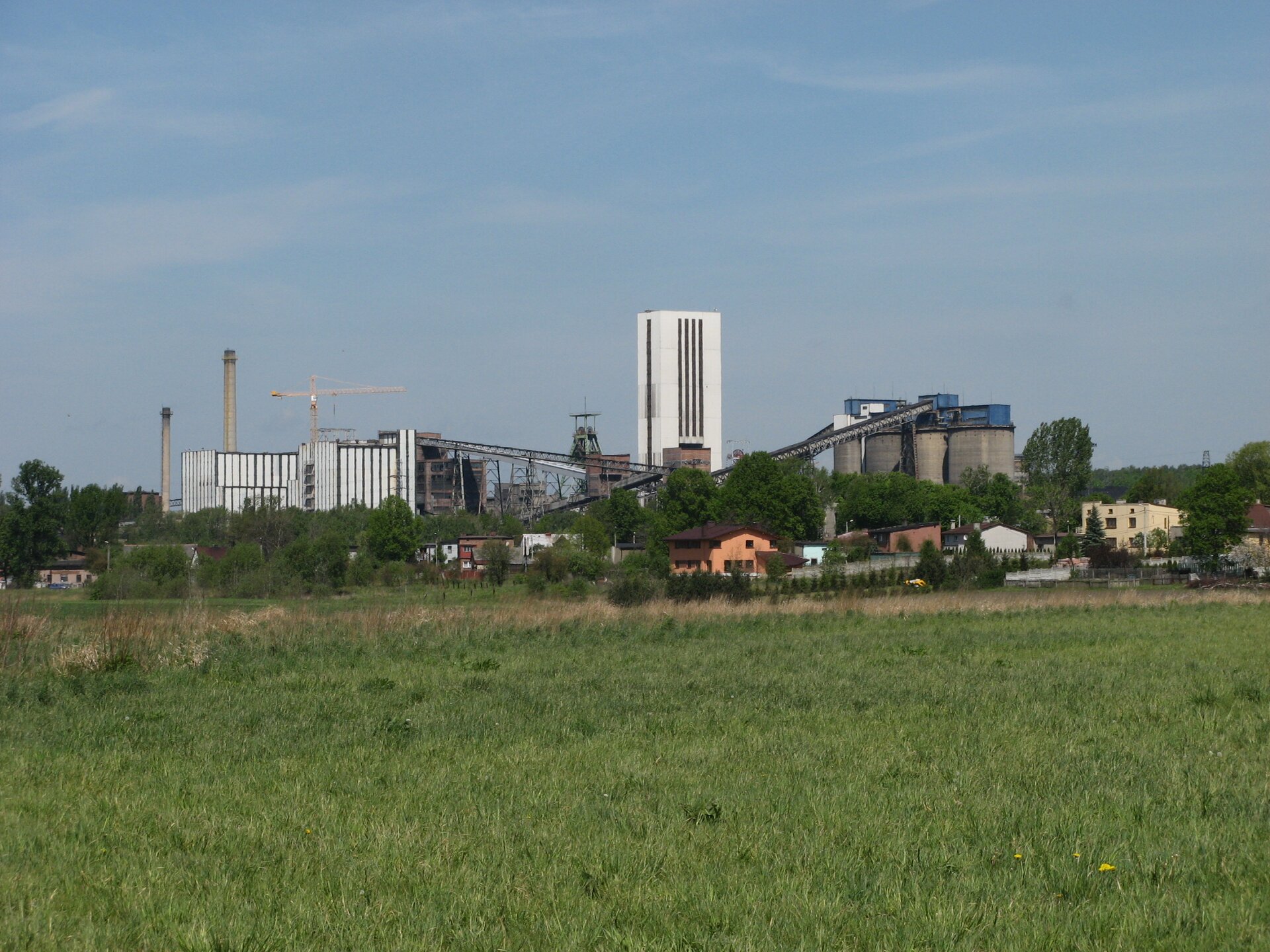 Zdjęcie przedstawia budynki kopalni węgla kamiennego Halemba w Rudzie Śląskiej. Słoneczny dzień, lato. Na pierwszym planie duża zielona polana. Na linii horyzontu, w planie średnim niskie budynki mieszkalne, a za nimi drzewa. Za drzewami budynki kopalni. W centrum zdjęcia, najwyższa budowla kopalni, biały budynek w kształcie pionowego prostopadłościanu. Jest to wieża wyciągowa. Na lewo dwa duże budynki, zakład przeróbczy. Ściany budynków pokryte pionowymi prostokątnymi białymi panelami. Za budynkami dwa wysokie okrągłe kominy wykonane z czerwonej cegły. Między kominami a wieżą wyciągową, znajduje się szyb górniczy wykonany z belek stalowych. Na szczycie szybu dwa koła wyciągowe. Po prawej stronie cztery wysokie budowle w kształcie walców o średnicy około dziesięciu metrów w szarym kolorze. To zbiorniki na węgiel. Wysokość budynków to połowa wysokości wieży wyciągowej. Dwa taśmociągi sięgają aż do dachu zbiorników na węgiel.