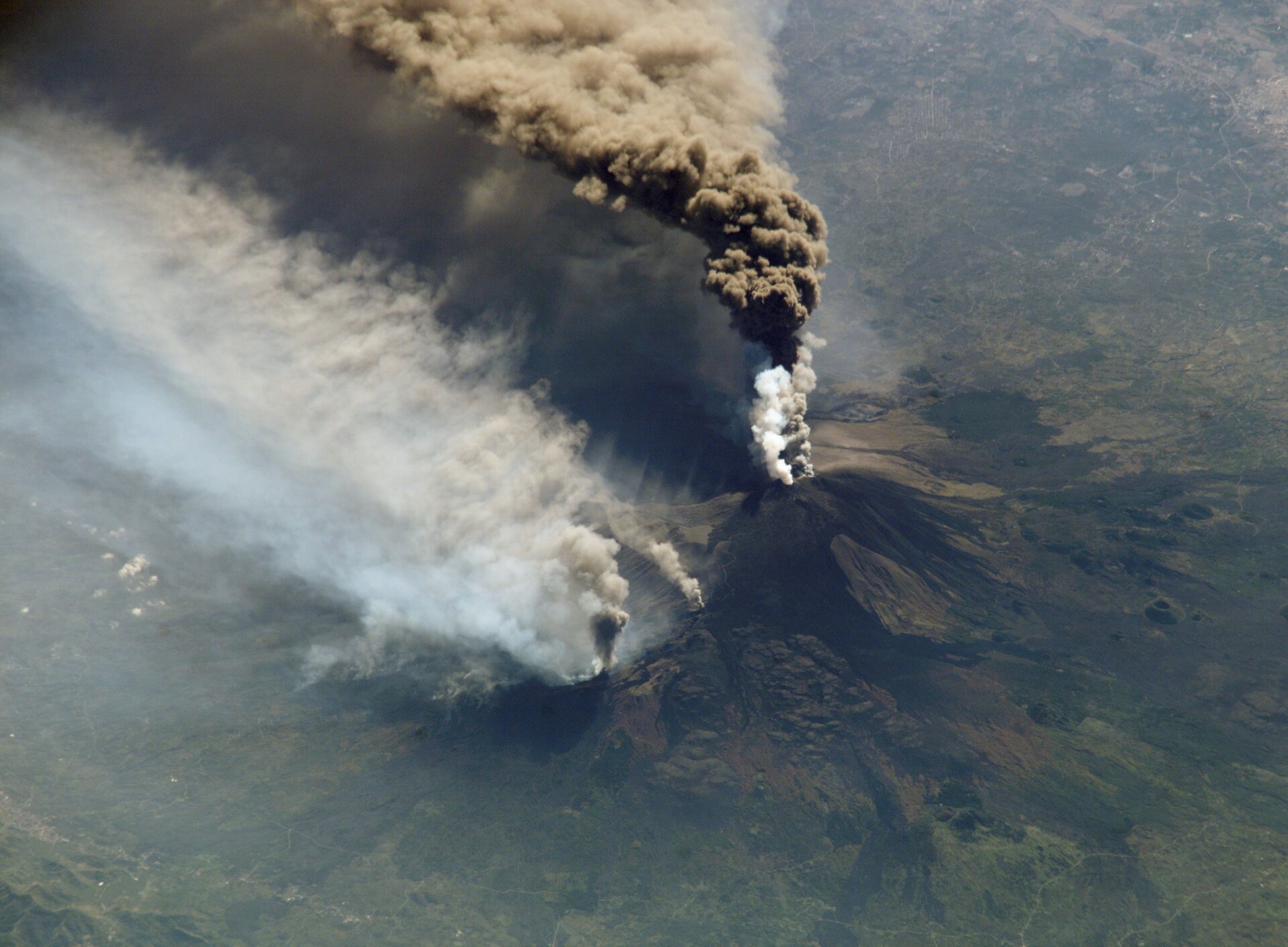 Fotografia wykonana z góry przedstawia wulkan podczas erupcji. Ze szczytu wulkanu wydobywa się chmura biało-brazowych gazów i pyłów.