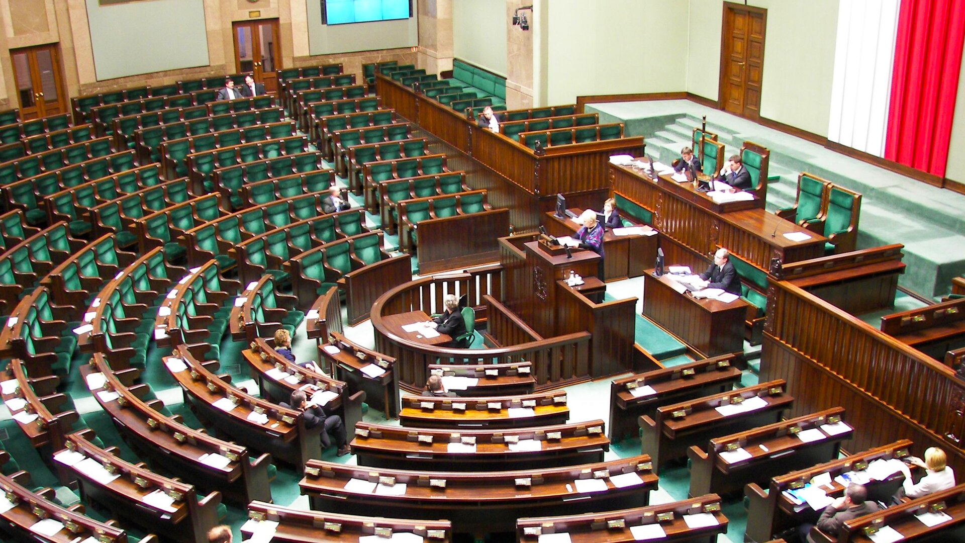 Ilustracja przedstawia salę posiedzeń w Sejmie. Na sali w półkolu umieszczone są dębowe siedzenia wyłożone zielonym materiałem, brązowe dębowe blaty i brązowa balustrada. U góry zdjęcia widać świecący na jasnoniebiesko fragment ekranu. Po prawej stronie widać brązowe drzwi na tle ściany w kolorze pistacjowym. Po środku ściany umieszczona jest od sufitu do podłogi biało-czerwona flaga Polski. Przed fotelami dla posłów umieszczona jest mównica, a za nią stoły dla Prezydium Sejmu. Na zdjęciu widać nielicznych posłów siedzących w swoich ławach poselskich, słuchających przemówienia pani poseł.