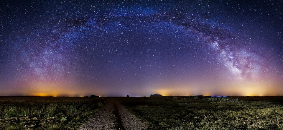 Zdjęcie przedstawiające panoramę nocną z odległymi światłami większego miasta. Na pierwszym planie znajduje się polna droga z nieużytkami po bokach. Droga prowadzi prosto w stronę horyzontu. Na niebie ogromna liczba gwiazd z wyraźnie odznaczającą się Drogą Mleczną tworzącą ogromny łuk nad całym kadrem.