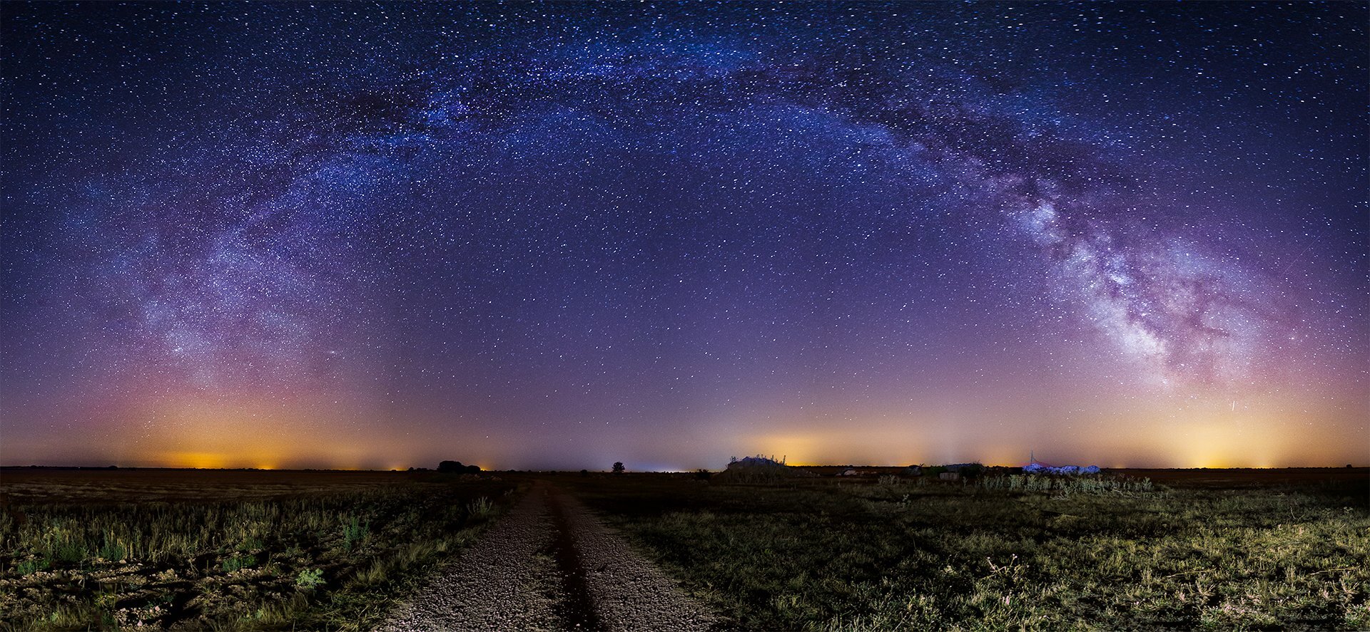 Zdjęcie przedstawiające panoramę nocną z odległymi światłami większego miasta. Na pierwszym planie znajduje się polna droga z nieużytkami po bokach. Droga prowadzi prosto w stronę horyzontu. Na niebie ogromna liczba gwiazd z wyraźnie odznaczającą się Drogą Mleczną tworzącą ogromny łuk nad całym kadrem.