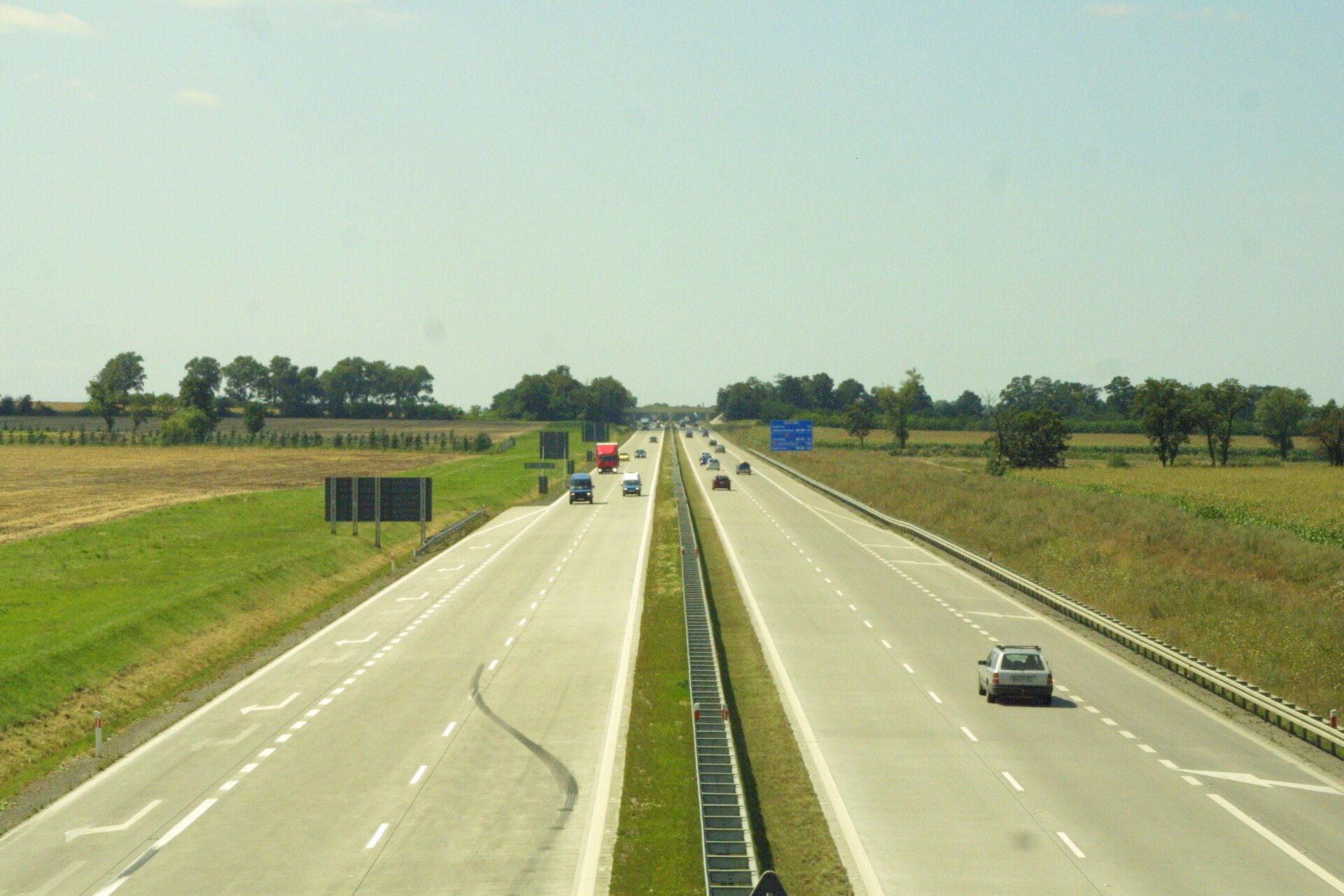 Na zdjęciu autostrada, dwa pasy i pobocze w każdym kierunku. Po obu stronach pola uprawne.