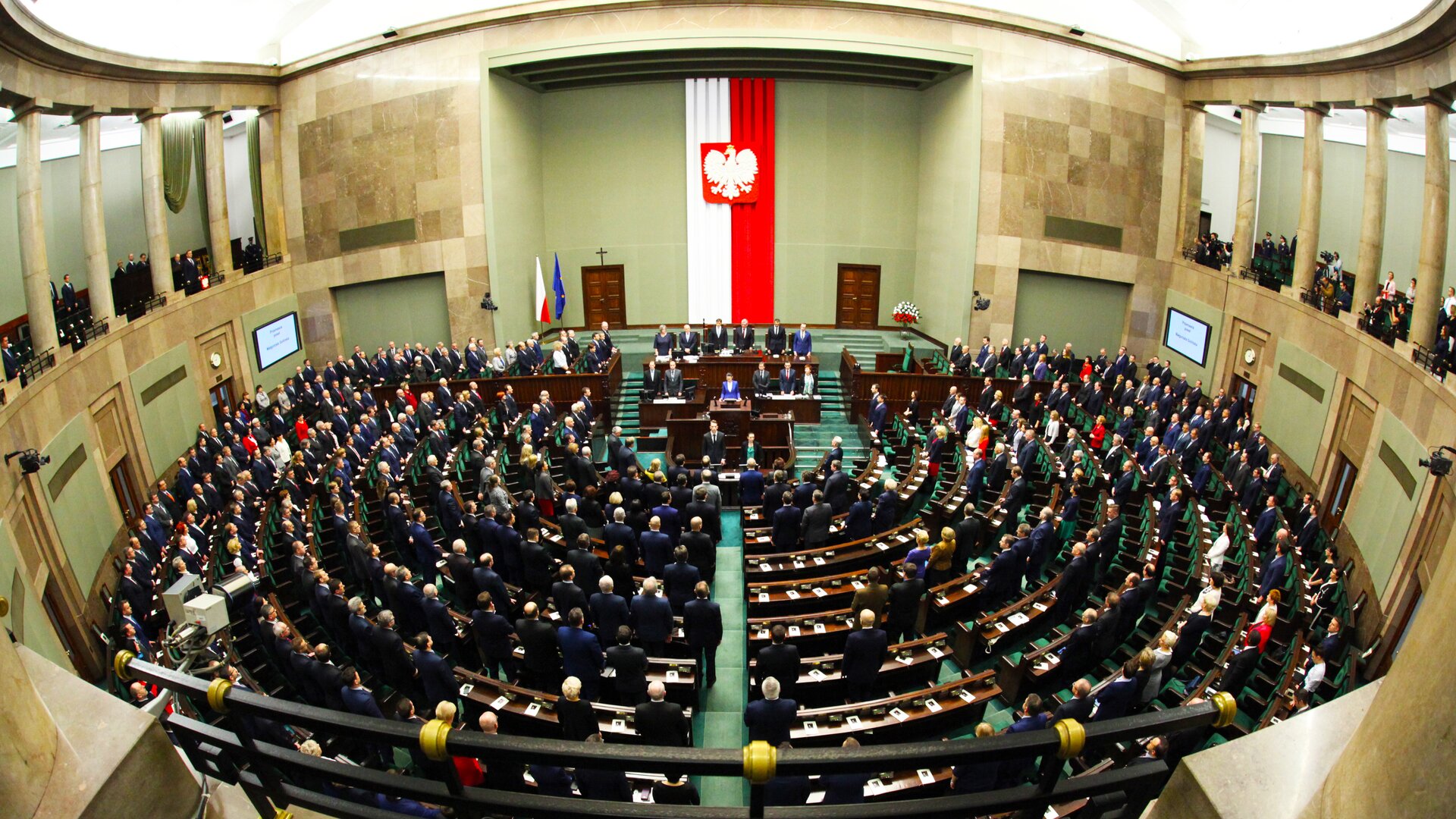 Na ilustracji znajduje się sala obrad Sejmu. W brązowych dębowych ławach stoją posłowie i posłanki. Sala jest całkowicie wypełniona. Zdjęcie sejmu jest zrobione z balkonu, na pierwszym planie widać balustradę. Po lewej i prawej stronie widać balkon sali sejmowej otoczony ozdobnymi kolumnami. W głębi ilustracji widać flagę Polską biało-czerwoną na tylnej ścianie. Po lewej stronie widzimy flagi na stojakach – flagę Unii Europejskiej, flagę Polski, drzwi, nad którymi jest umieszczony krzyż. Po prawej stronie stoi w wazonie bukiet z kwiatów biało-czerwonych.