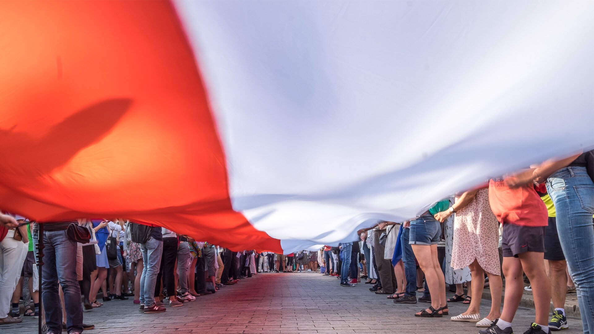 Ilustracja przedstawia zdjęcie bardzo licznej grupy ludzi uczestniczących w patriotycznej manifestacji. Duża grupa uczestników ustawionych w dwa szeregi ludzi ( kobiety i mężczyźni ) trzyma rozciągniętą w poziomie bardzo długą biało-czerwoną flagę Polski. Ujęcie jest bardzo ciekawe. Zdjęcie zrobione jest „ od spodu”, z poziomu powierzchni ulicy. Rozciągnięta flaga przesłania niebo i sylwetki od pasa w górę trzymających ją ludzi. Poza osobami trzymającymi flagę plac, na którym odbywa się manifestacja, wypełnia tłum innych uczestników manifestacji.