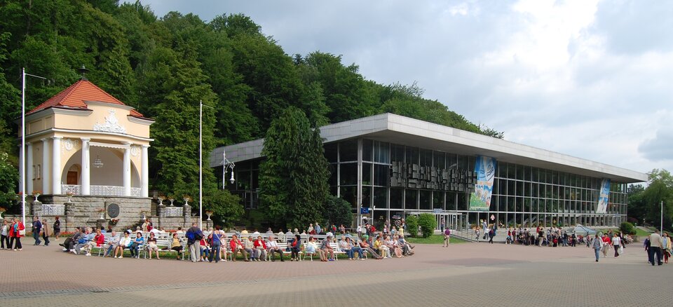 Na zdjęciu nowoczesny oszklony budynek z plaskim dachem. Przed budynkiem deptak, ławki, ludzie.