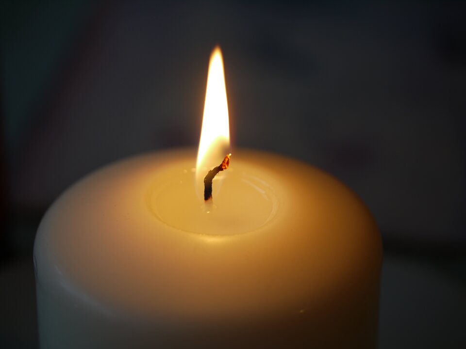 Zdjęcie przedstawia górną część świecy. Świeca ma średnicę około pięć centymetrów. Na szczycie świecy krótki knot. Knot jest zapalony. Koniec knota jest rozżarzony i czerwony. Dolna część knota jest czarna. Koniec knota wychodzący ze świecy jest biały. Cały knot od części czarnej do czerwonej jest w płomieniu. Płomień ma kolor biały. Płomień ma kształt wydłużonego trójkąta. Tło zdjęcia czarne.