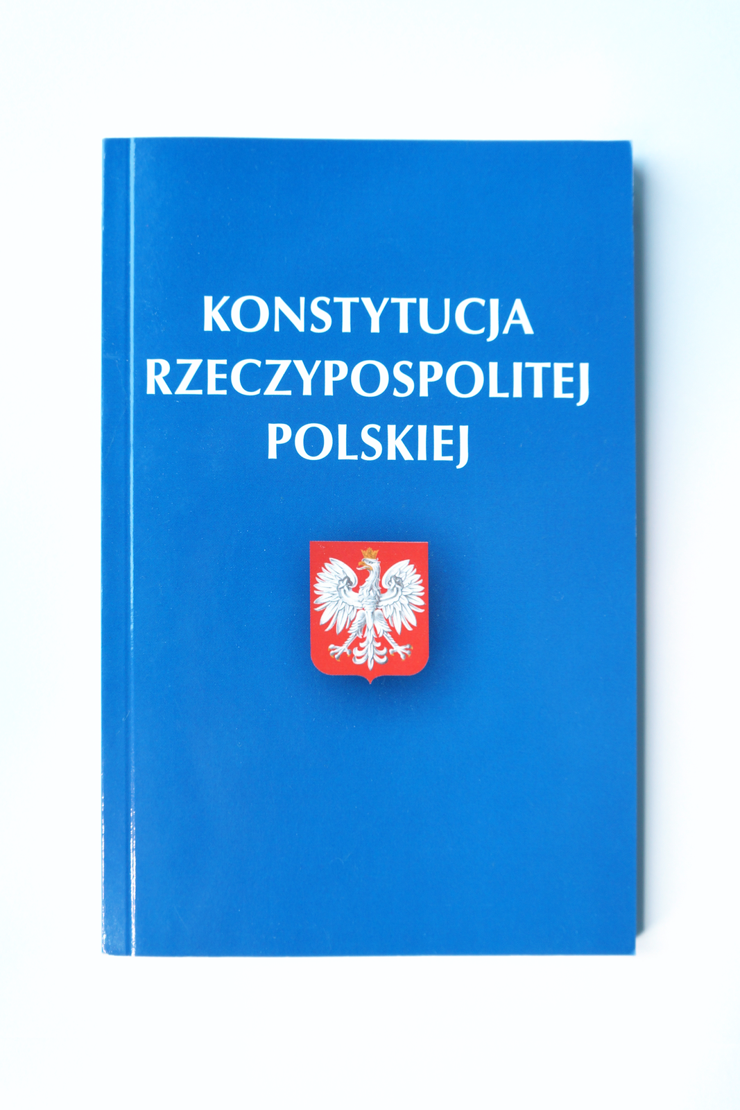 Ilustracja przedstawia prostokątną okładkę książki, na której umieszczono biały napis Konstytucja Rzeczypospolitej Polskiej. Poniżej godło Polski – biały orzeł na czerwonym tle. Głowa orła skierowana jest w prawo.