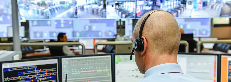 Zdjęcie przedstawia centrum monitorujące ważne miejsca podlegające kontroli. Centrum to pomieszczenie biurowe. Na pierwszym planie, odwrócony tyłem, siedzi przy biurku funkcjonariusz centrum zarządzania. Przed nim ustawionych jest kilka monitorów. Naprzeciwko, w głębi centrum, ekran pokrywający całą ścianę. Ekran podzielony jest na mniejsze monitory. Na monitorach miejsca takie jak wejścia do metra, plany miasta.