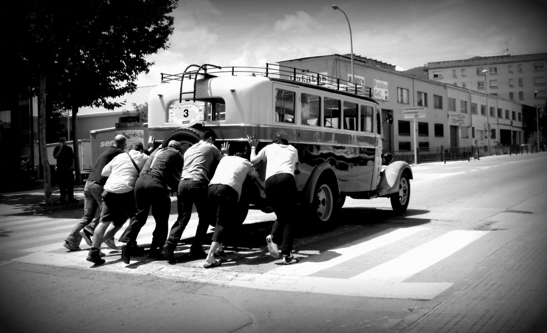 Czarno białe zdjęcie przedstawiające sześciu mężczyzn przepychających stary autobus ulicami małego miasteczka. Na budynkach w głębi kadru widoczne są hiszpańskojęzyczne napisy.