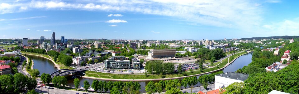 Na pierwszym planie rzeka, a za nią szeroka panorama nowoczesnego, dużego miasta. Z prawej stronie grupa wieżowców górująca nad reszta miasta.