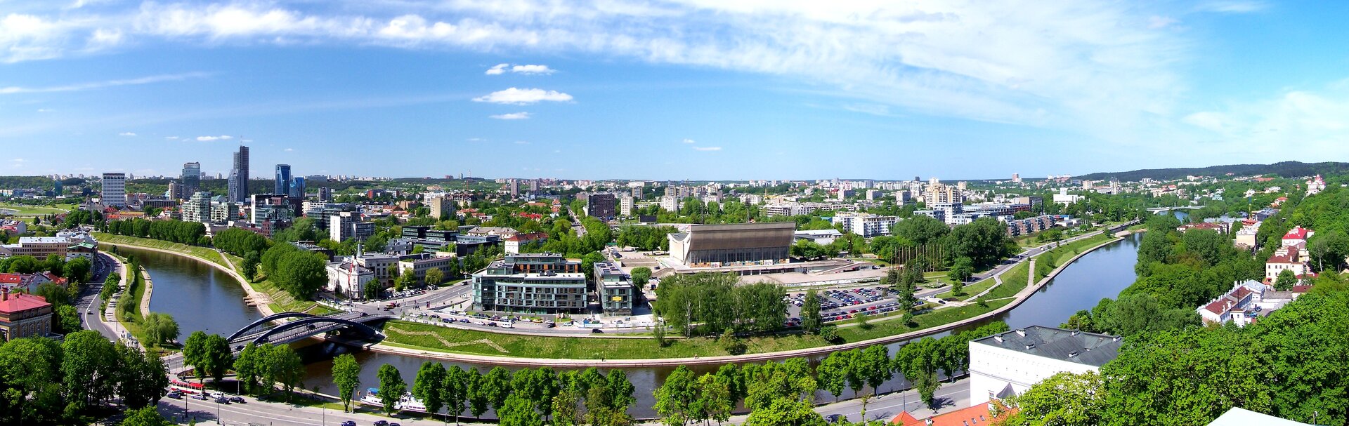 Na pierwszym planie rzeka, a za nią szeroka panorama nowoczesnego, dużego miasta. Z prawej stronie grupa wieżowców górująca nad reszta miasta.
