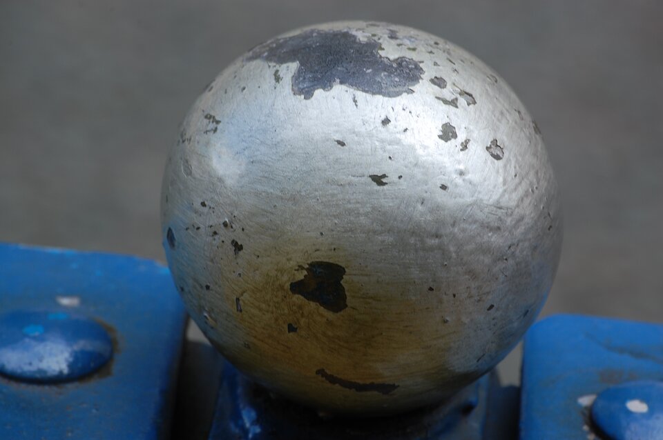 Zdjęcie przedstawia małą metalową kulkę z odpryskami srebrnej farby w kilku miejscach na powierzchni. Kulka stoi na jakiejś metalowej podstawie pomalowanej na niebiesko, która może być z kolei elementem konstrukcyjnym jakiegoś mostu lub poręczy.