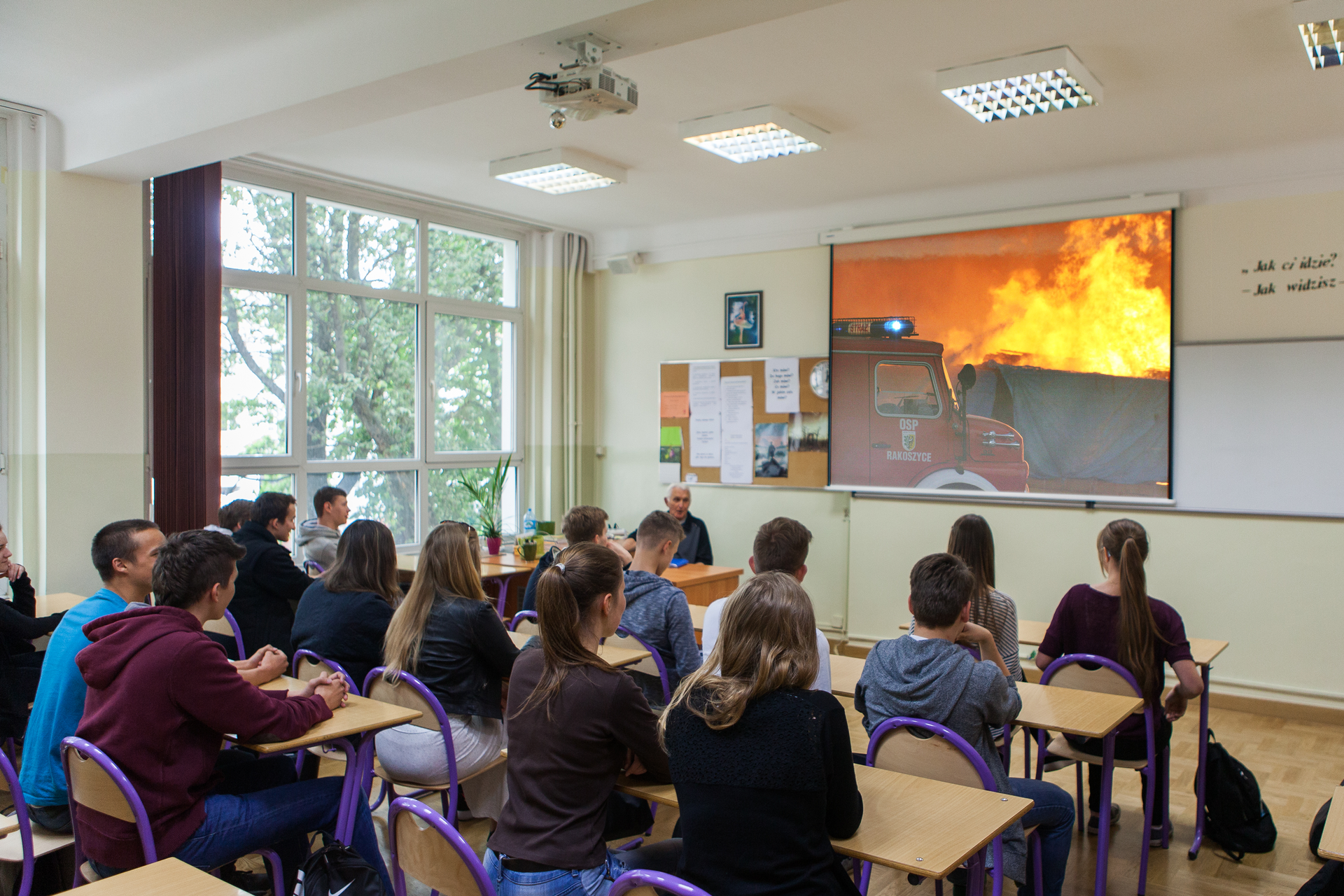 Kolorowe zdjęcie klasy lekcyjnej. Grupa uczniów siedzi w ławkach tyłem do obserwatora zdjęcia, przodem do tablicy. Na ścianie, obok tablicy, rozciągnięty ekran. Na ekranie fragment filmu dotyczący ochrony przeciwpożarowej. Film przedstawia straż pożarną gaszącą pożar. Nauczyciel siedzi za biurkiem, w lewym rogu klasy.