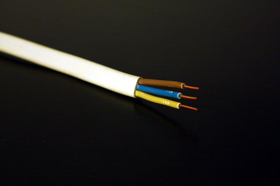Fotografia przedstawia kabel elektryczny. Kabel biały. Tło ciemne, prawie czarne. Kabel został przecięty. Widać 3 przewody, które znajdują się wewnątrz. W środku każdego znajduje się miedziany drut. Przewody mają różne kolory: brązowy, niebieski i żółty.