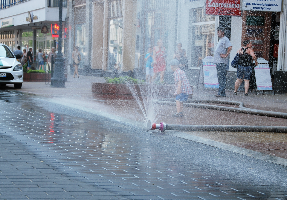 Zdjęcie w kształcie poziomego prostokąta. Dzień. Na pierwszym planie ulica i chodnik w centrum miasta pokryte kostką brukową. Po prawej wąż strażacki. Wąż rozciągnięty poprzecznie przez chodnik aż do krawężnika. Koniec węża zakończony zaworem. Woda tryskająca pod dużym ciśnieniem, tworzy strumień w kształcie wachlarza. Jest to tak zwana kurtyna wodna. Za strumieniem wody mały chłopiec w czapce. Za chłopcem przechodnie. Osoby ubrane w lekkie letnie ubrania. W tle sklepy, latarnie i kwietniki.