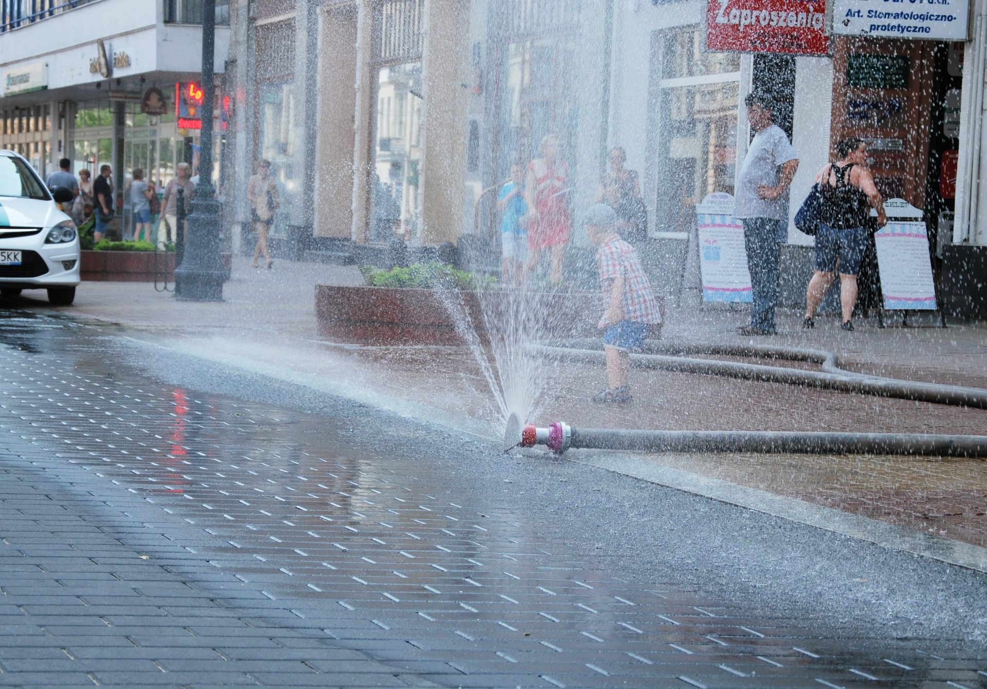 Zdjęcie w kształcie poziomego prostokąta. Dzień. Na pierwszym planie ulica i chodnik w centrum miasta pokryte kostką brukową. Po prawej wąż strażacki. Wąż rozciągnięty poprzecznie przez chodnik aż do krawężnika. Koniec węża zakończony zaworem. Woda tryskająca pod dużym ciśnieniem, tworzy strumień w kształcie wachlarza. Jest to tak zwana kurtyna wodna. Za strumieniem wody mały chłopiec w czapce. Za chłopcem przechodnie. Osoby ubrane w lekkie letnie ubrania. W tle sklepy, latarnie i kwietniki.