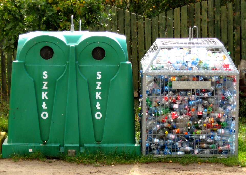 Na zdjęciu pojemniki do recyclingu. Z lewej strony zielony pojemnik z okrągłym otworem w górnej części. Napis szkło. Z prawej strony pojemnik z siatki. Wewnątrz plastikowe butelki.