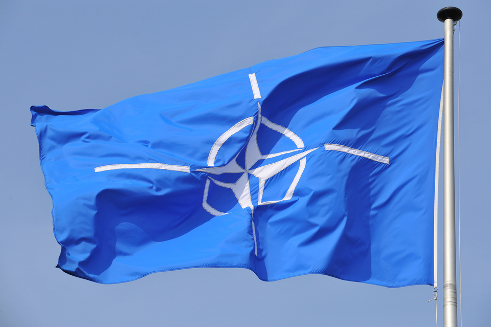 Zdjęcie przedstawia flagę NATO zawieszoną na maszcie. Maszt po prawej stronie. Flaga unosi się i jest skierowana w lewo. Na błękitnym tle znajduje się biały pierścień. W środku pierścienia czteroramienna gwiazda. To tak zwana róża kompasowa. Ramiona gwiazdy są zakończone białym, wąskim paskiem.