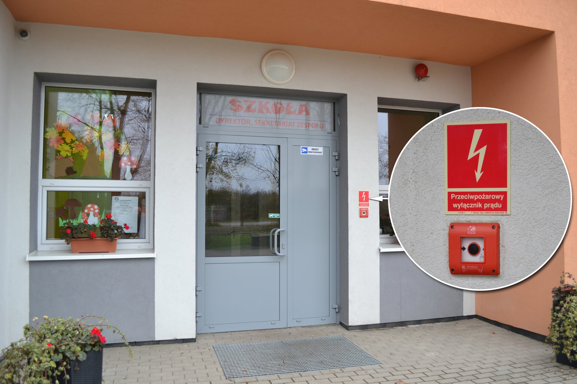 Kolorowe ujęcie głównego wejścia do budynku szkoły za dnia. Wejście to duże przeszklone pojedyncze drzwi z czerwonym napisem szkoła powyżej. Z prawej strony drzwi mieście się przeciwpożarowy wyłącznik prądu, który sygnalizuje czerwona ramka. Powyżej czerwona prostokątna tabliczka. Tabliczka podzielona na dwie części. Górna część, zajmująca dwie trzecie tabliczki, to czerwony prostokąt. Przez środek prostokąta przebiega biała błyskawica w kształcie strzałki z grotem skierowanym ku dołowi. W połowie strzałka jest zakrzywiona w formie zygzaka. Dół tabliczki to biały napis: główny wyłącznik prądu.