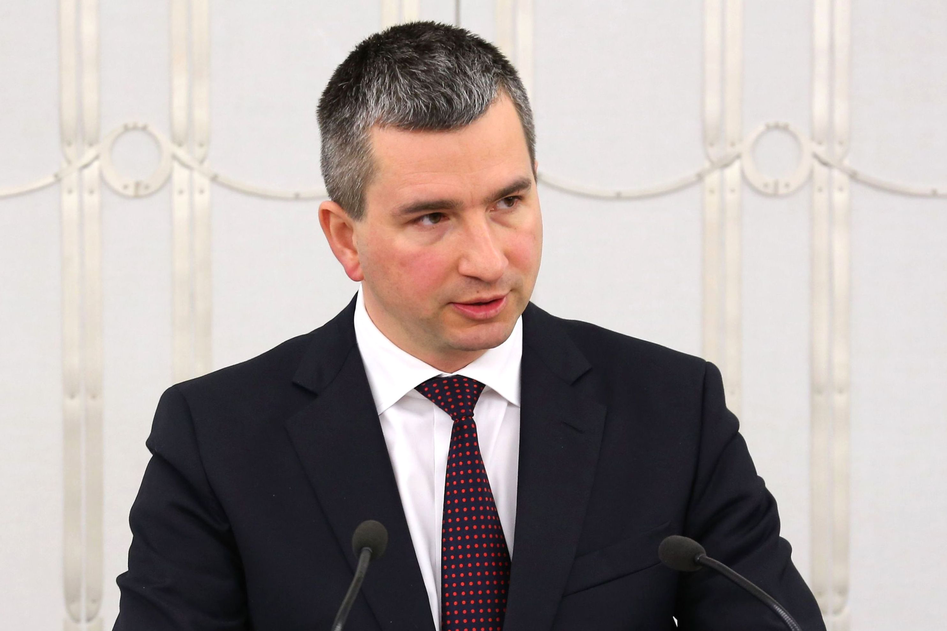 Kolorowe zdjęcia portretowe Mateusza Szczurka. Mężczyzna przodem do obserwatora zdjęcia. Wiek, około 45 lat. Czarne włosy z siwymi kosmykami zaczesane do przodu. Włosy krótkie, gęste, z wyraźnymi zakolami nad czołem. Czoło wysokie, gładkie. Brwi ciemne, gęste. Oczy ciemne, małe. Nos duży. Usta wąskie. Twarz okrągła. Policzki pulchne. Uszy duże, przylegające. Ubrany w czarną marynarkę, białą koszulę i bordowy wzorzysty krawat.