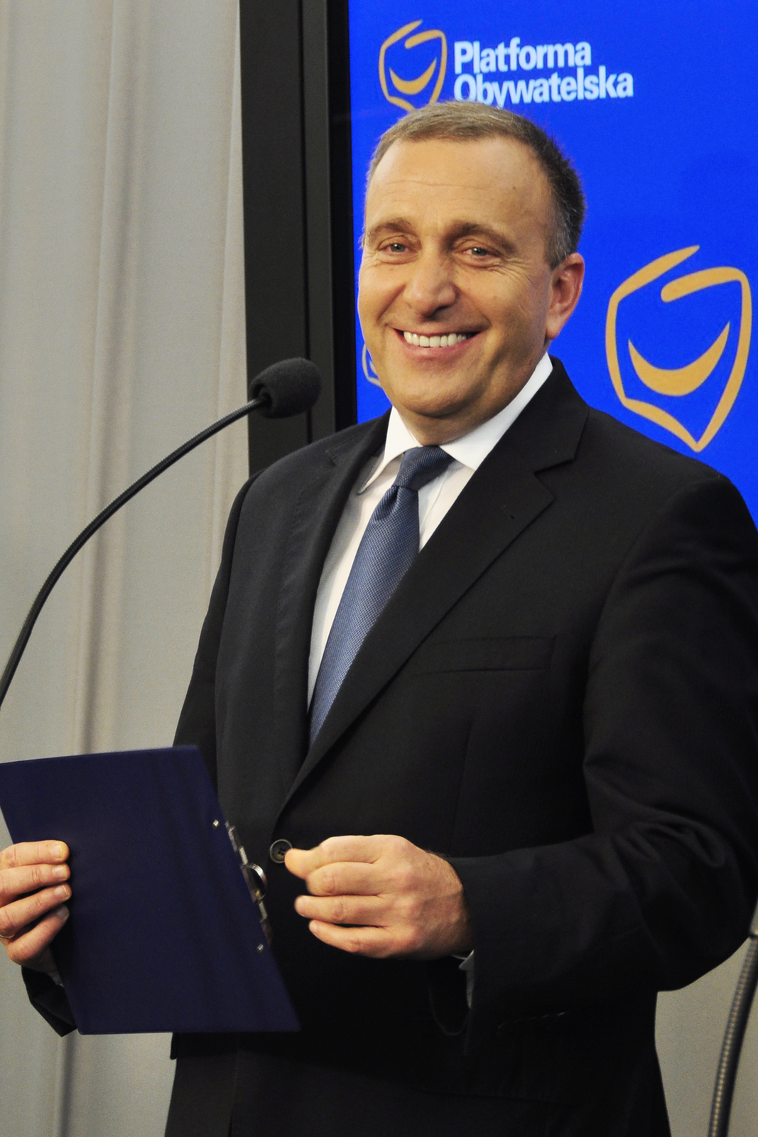 Zdjęcie przedstawia Grzegorza Schetynę. Mężczyzna stoi przed mikrofonem. Mężczyzna w wieku około 50 lat. Twarz podłużna, gładko ogolona. Włosy ciemne, krótkie, zaczesane gładko. Czoło wysokie. Brwi ciemne, gęste. Oczy małe. Nos duży. Usta wąskie. Mężczyzna uśmiecha się. Uszy przylegające. Mężczyzna ubrany w czarną marynarkę, białą koszulę i niebieski krawat. Mężczyzna trzyma przed sobą granatową plastikową podkładkę.