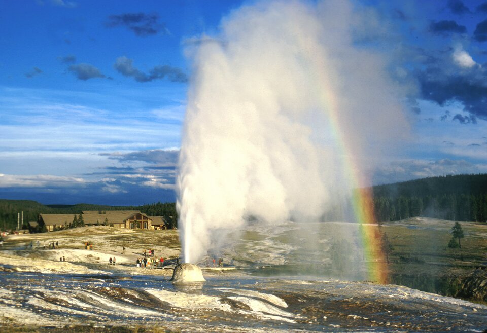 Fotografia przedstawia tryskający wodą gejzer w Parku Narodowym Yellowstone. Z boku w kropach wody utworzyła się tęcza. Gejzer to wypukłość na szaro – brązowo – białym podłożu. Wysokość wytrysku wody można ocenić, porównując ją z malutkimi postaciami ludzi na dalszym planie. W tle zabudowania i las.