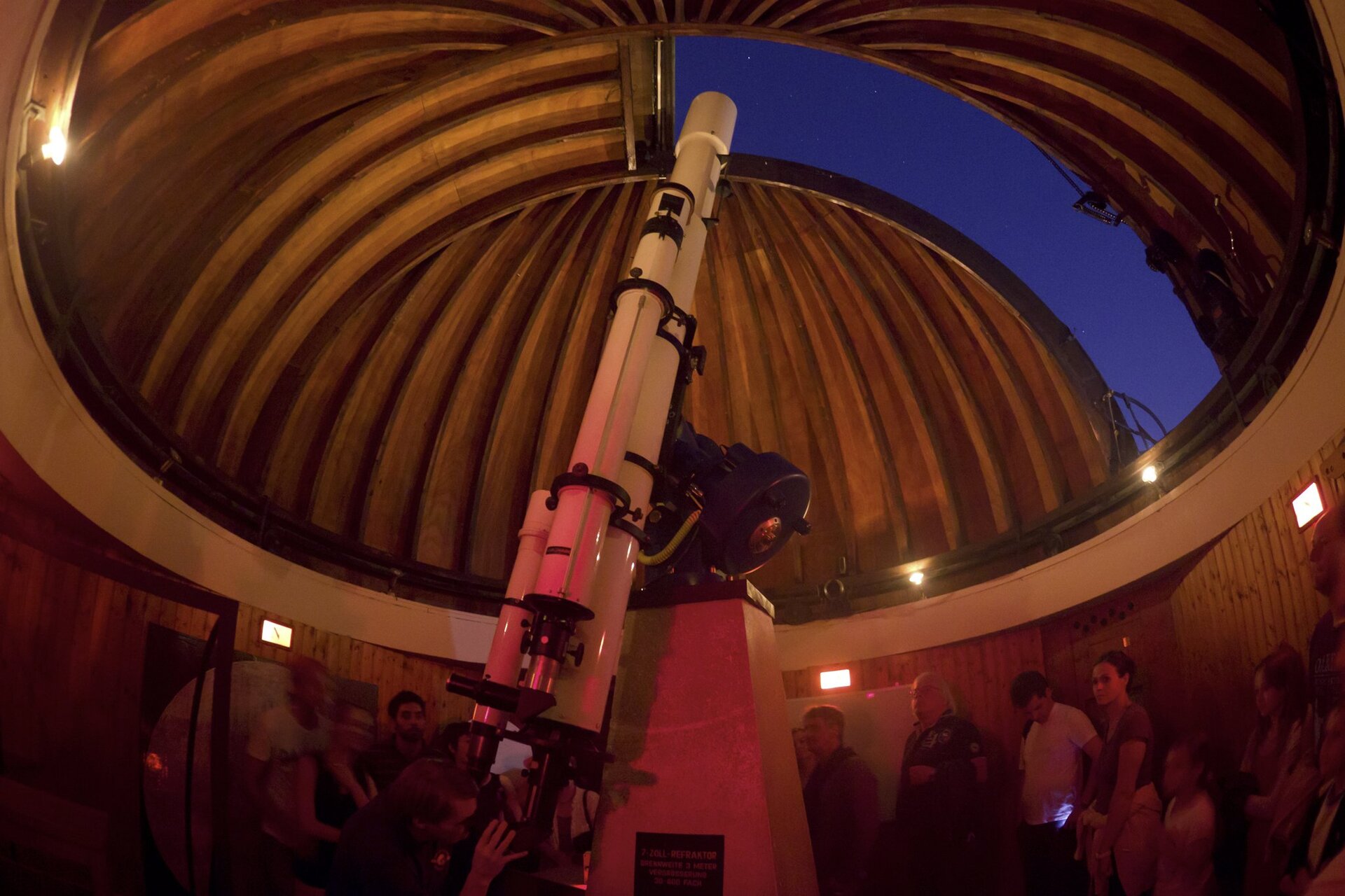 Zdjęcie przedstawia wnętrze obserwatorium w Monachium podczas pokazowej lekcji astronomii. W kadrze znajduje się długi, ustawiony w pozycji pionowej biały teleskop i otwarta kopuła z widocznym nocnym niebem. Poniżej wokół teleskopu zebrani ludzie w różnym wieku. Młody mężczyzna nachylony przed teleskopem spogląda przez okular.