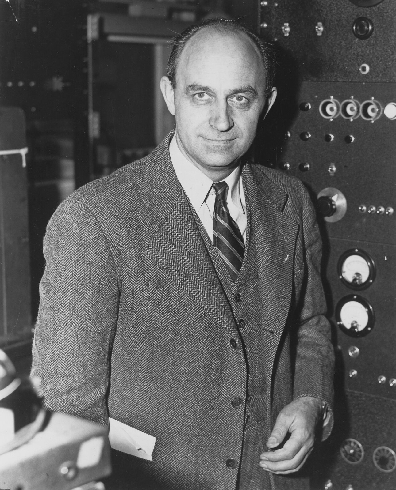 Zdjęcie przedstawia Enrico Fermi. Fotografia czarno-biała. Na zdjęciu mężczyzna w wieku ok. 45 lat, szczupły, łysiejący. Czoło wysokie, linia włosów cofnięta, włosy ciemne. Brwi gęste. Oczy duże. Nos długi. Usta wąskie. Mężczyzna się uśmiecha. Widoczne cienie pod oczami. Uszy duże, delikatnie odstające. Mężczyzna jest ubrany w trzyczęściowy garnitur w prążki, białą koszulę oraz krawat w paski.
