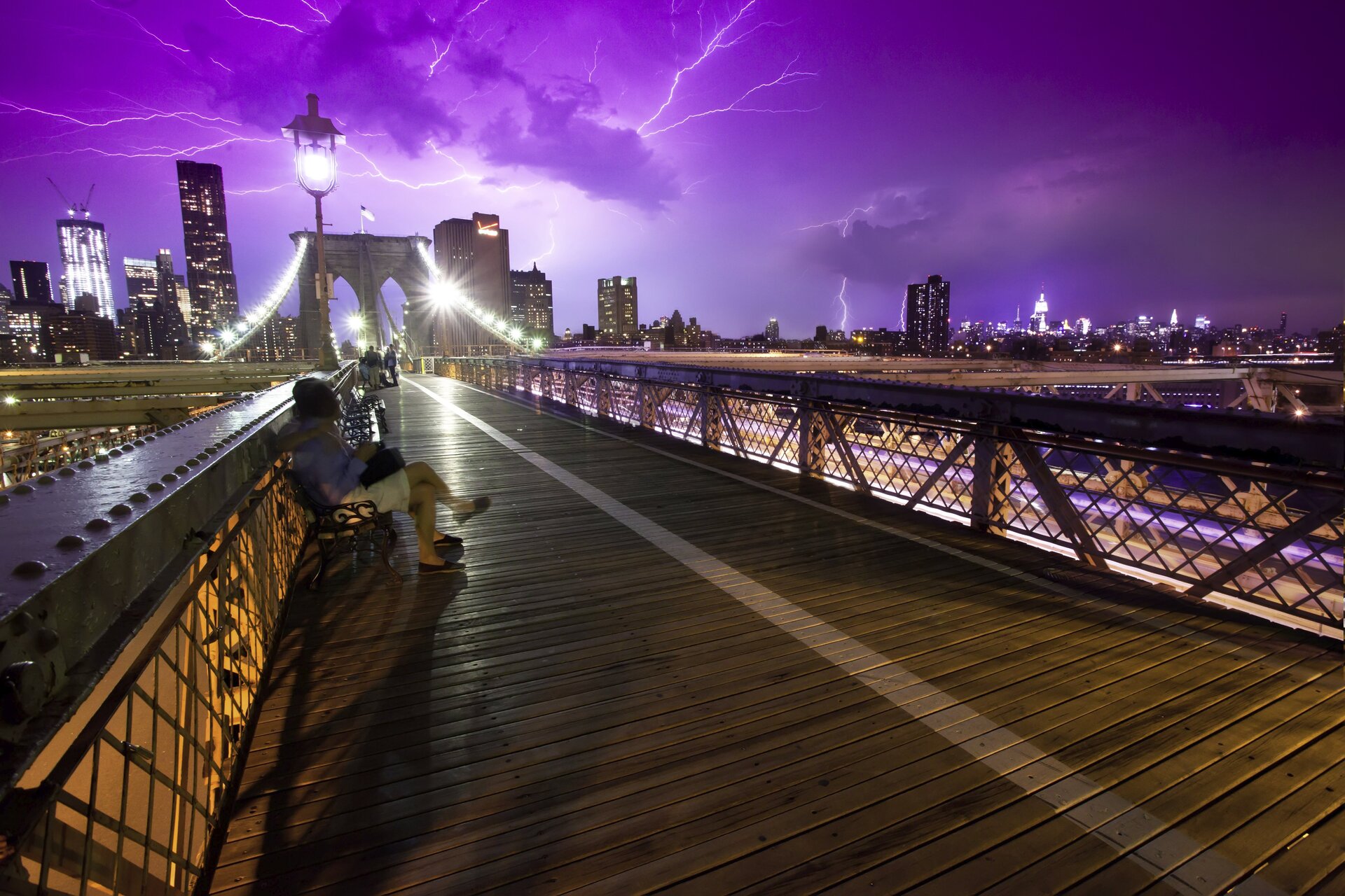 Zdjęcie przedstawia niecodzienną scenę. Noc, panorama Nowego Jorku z mostem brooklyńskim w centrum kadru. Daleko w tle światła Manhattanu, na pierwszym planie pomost z ławkami na przedłużeniu którego w oddali znajduje się centralna część mostu. Pierwsza ławka zajęta przez siedzącą na niej kobietę. Widoczne światła wielkiego miasta oraz oświetlenie mostu i latarnie przy kładce. Niebo pochmurne w kolorze fioletu, całe poprzecinane błyskawicami odległej burzy.
