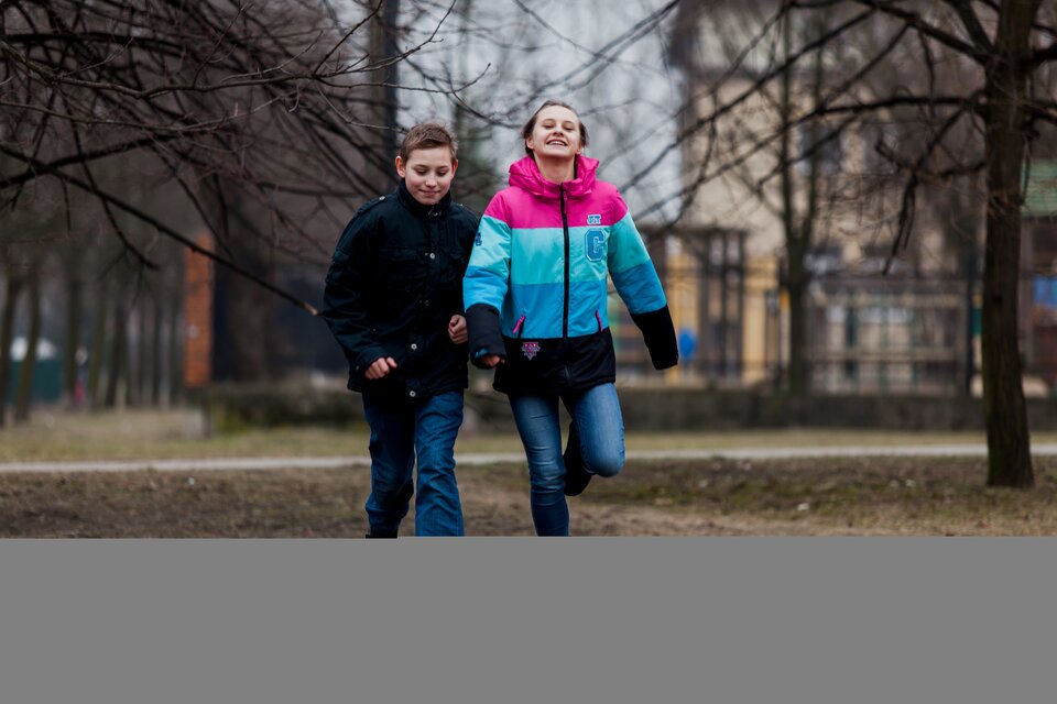Fotografia przedstawia boisko do gry w piłkę nożną. Na pierwszym planie dwóch chłopców kopiących piłkę. W tle widoczni inni chłopcy w strojach sportowych wykonujący ćwiczenia fizyczne