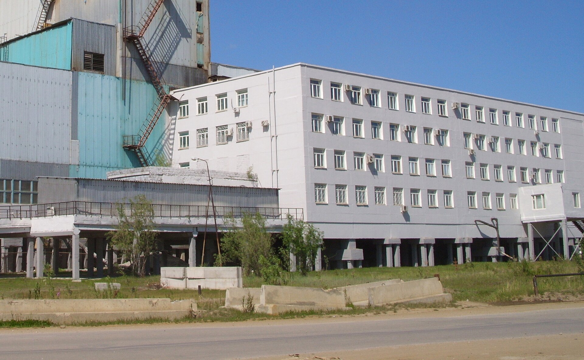 Fotografia prezentuje trzypiętrowy, biały, betonowy budynek z wieloma oknami. Budynek wzniesiono na betonowych palach wbitych w ziemię.