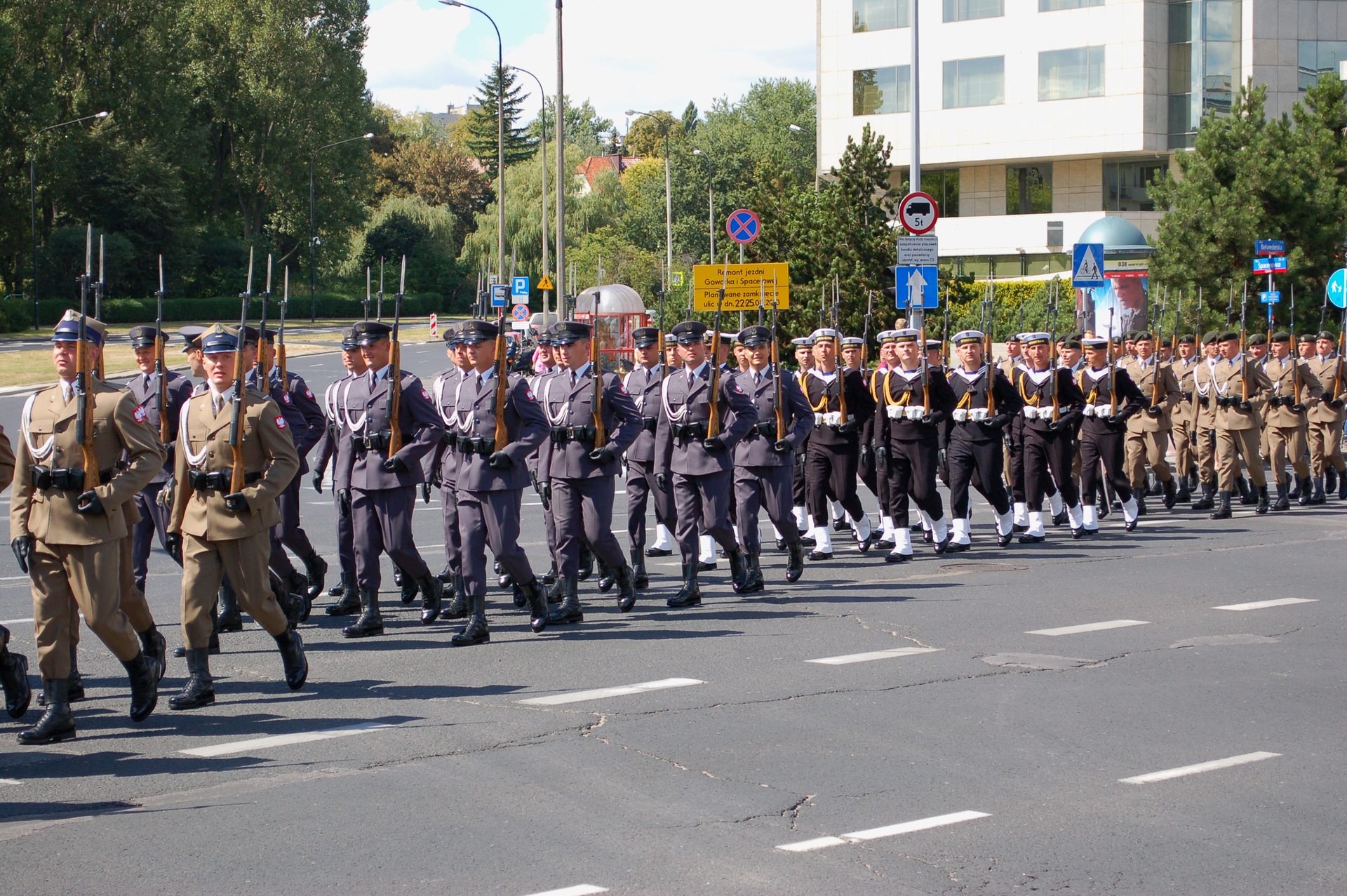 Kolorowe zdjęcie przedstawia defiladę sił zbrojnych RP. Słoneczny letni dzień. Ulica. Od prawej strony zdjęcia, w lewo, kolumna żołnierzy maszerujących w defiladzie. Wśród żołnierzy grupy różnych formacji. Od prawej strony zdjęcia: żołnierze wojsk pancernych w zielonych beretach i jasno-brązowych mundurach. Na lewo żołnierze marynarki wojennej w czarnych mundurach, białych rękawiczkach, i białych czapkach. W centralnej części zdjęcia żołnierze wojsk lotniczych w szarych mundurach. Po lewej stronie zdjęcia żołnierze wojsk lądowych. Każdy żołnierz trzyma karabin w lewej ręce. Karabiny skierowane pionowo w górę, wsparte kolbą na dłoni.