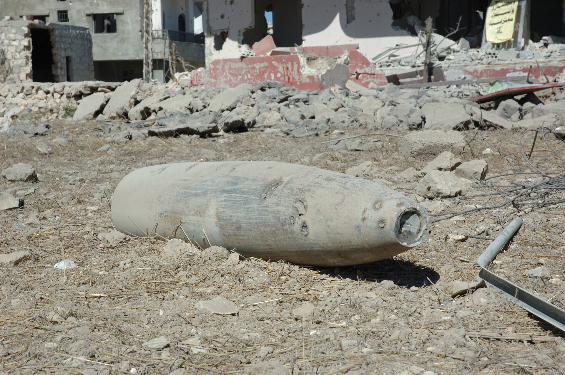 Fotografia prezentuje dużych rozmiarów pocisk leżący na ziemi , dookoła gruzy budynku