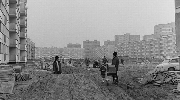 Fotografia przedstawia plac budowy wielopiętrowych bloków z wielkiej płyty. Na plac budowy jest piach, materiały budowlane a pomiędzy nimi mieszkańcy spacerujący z dziećmi.