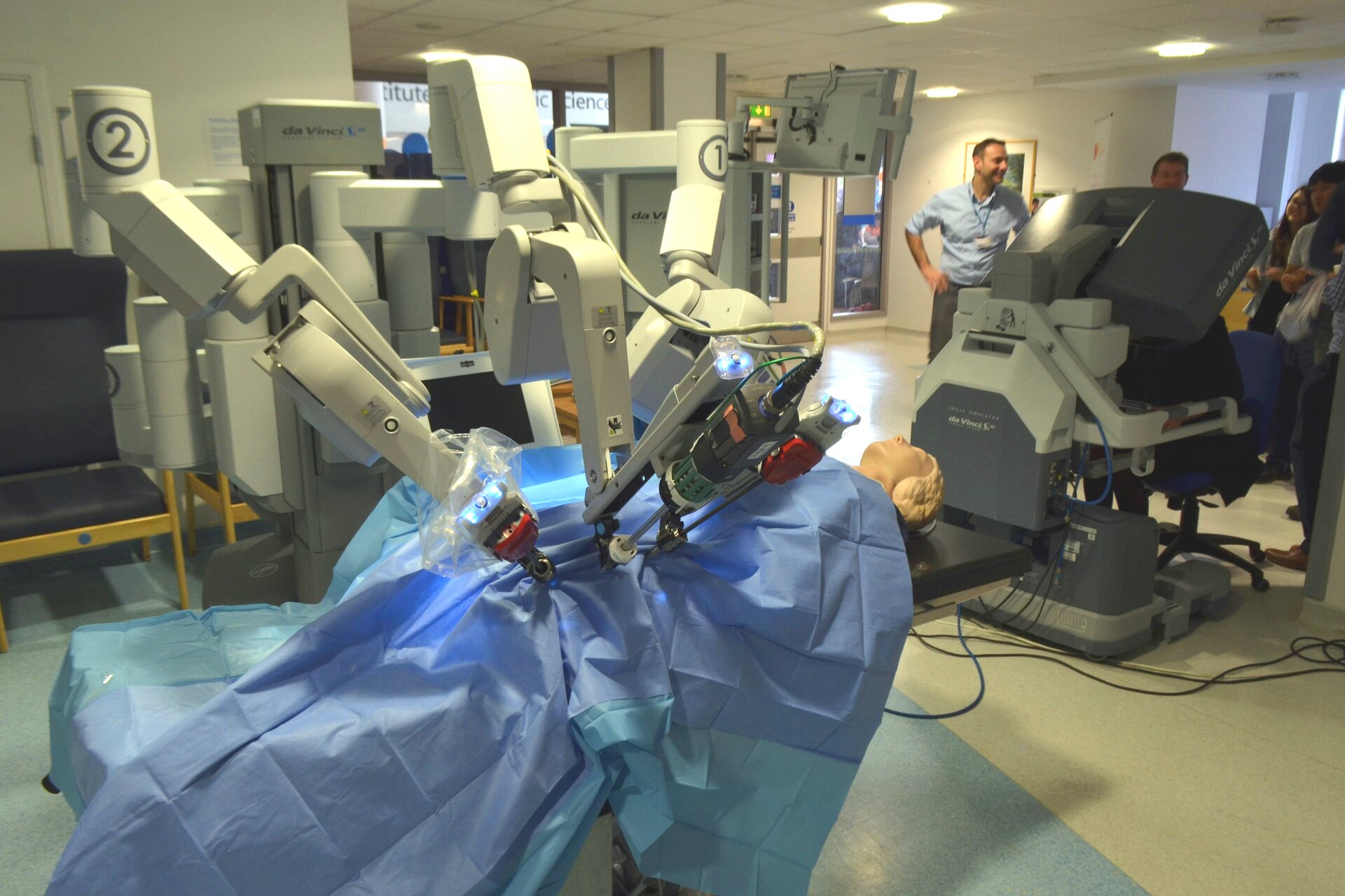 Na zdjęciu system robota medycznego. System składa się z dwóch odrębnych części – konsoli chirurgicznej oraz robota medycznego, posiadającego kilka ramion. Przy konsoli stoją ludzie. Na łóżku operacyjnym leży manekin, przykryty niebieskim prześcieradłem. W tle inne urządzenia medyczne.