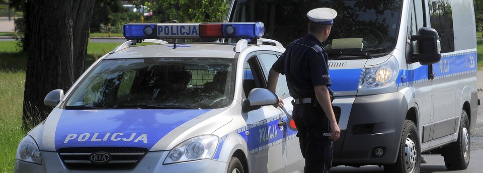 Kolorowe zdjęcia przedstawia dwa wozy policyjne zwrócone przodem do obserwatora. Tyłem do obserwatora, przy pierwszym wozie stoi policjant, ubrany w mundur z krótkim rękawem. Policjant patrzy w stronę nadjeżdżających samochodów.