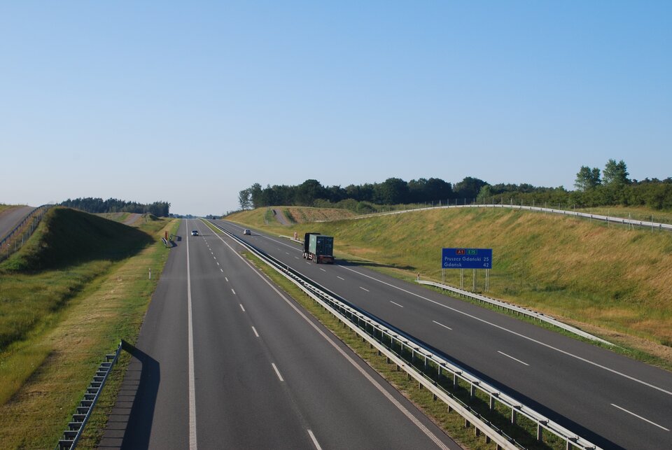 Na zdjęciu prosty odcinek autostrady, dwa pasy drogi i pobocze w każdym kierunku, ogrodzona siatką.