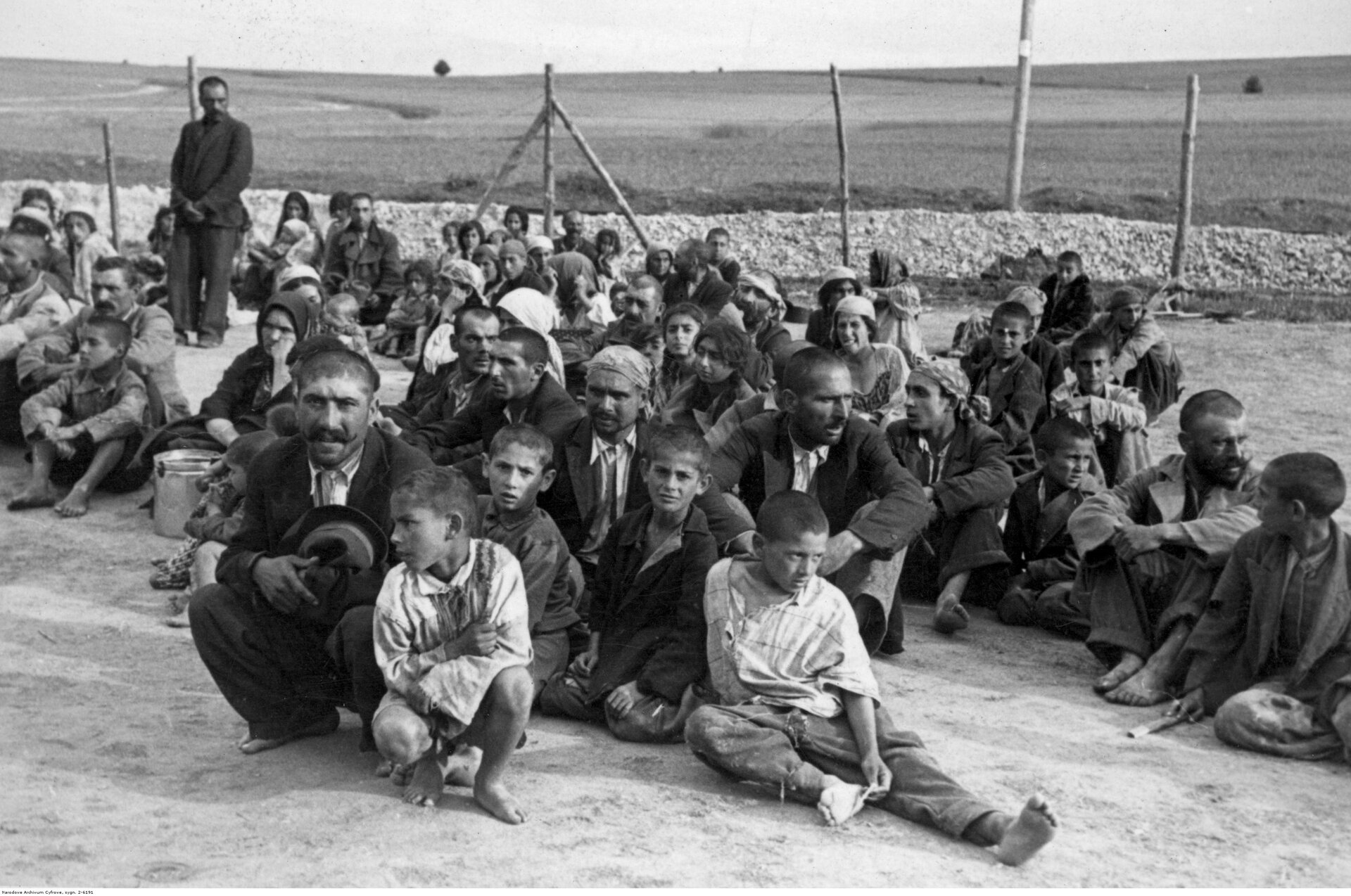 Stara fotografia. Na zdjęciu grupa Cyganów siedzących na ziemi, dorośli i dzieci. Za nimi ogrodzenie. Jeden mężczyzna stoi przy ogrodzeniu.