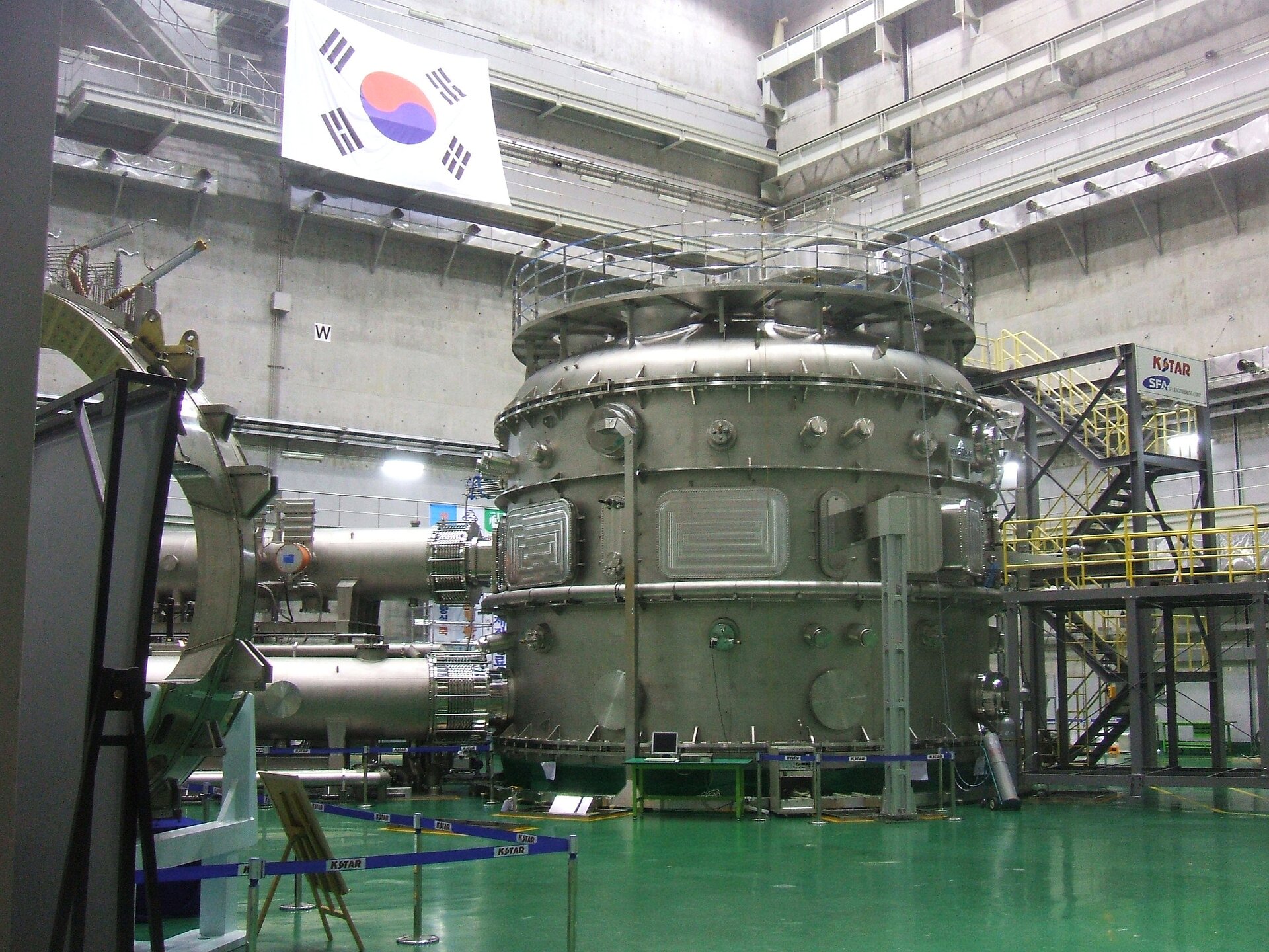 Zdjęcie przedstawia tokamak w Korei Południowej stojący w dużym pomieszczeniu fabrycznym o zielonej podłodze. Wysoko na platformie obserwacyjnej wisi flaga Korei. Sam tokamak ma postać dużego, beczkowatego urządzenia, od którego z lewej strony odchodzi system rur. Rusztowania wokół pozwalają ocenić wysokość maszyny na co najmniej osiem metrów.