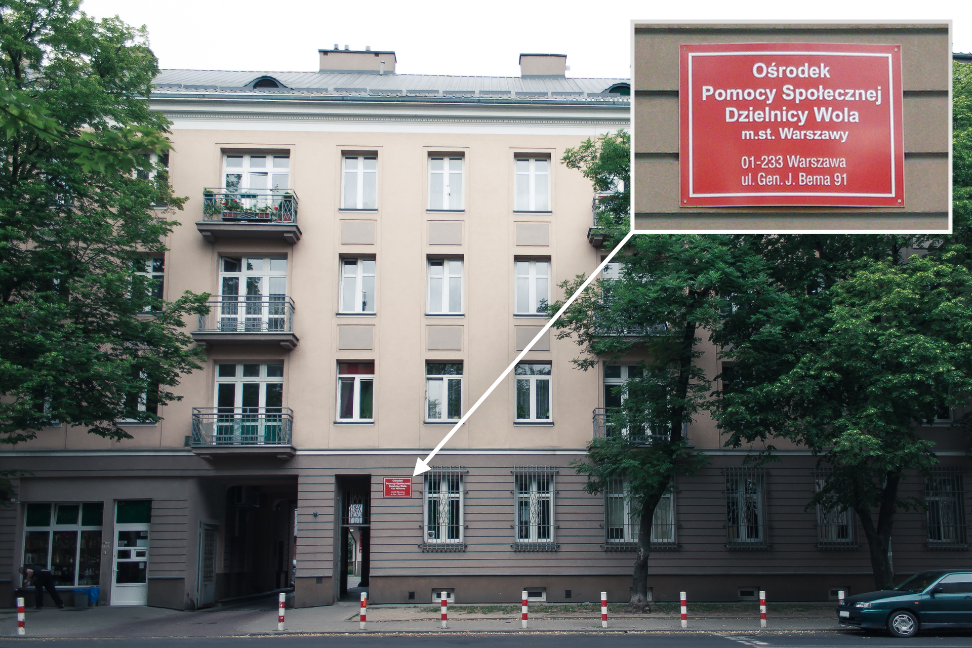 Kolorowe zdjęcie przedstawia duży trzypiętrowy budynek. Ściana brązowa na wysokości parteru. Powyżej parteru ściana w kolorze beżowym. W środkowej części brama wjazdowa prowadząca na dziedziniec budynku. Po prawej drzwi. Powyżej duża czerwona tablica z informacją: Ośrodek Pomocy Społecznej Dzielnicy Wola m.st. Warszawy. Warszawa. ul. Gen. J. Bema 51. Tablica powiększona w prawym górnym rogu zdjęcia.