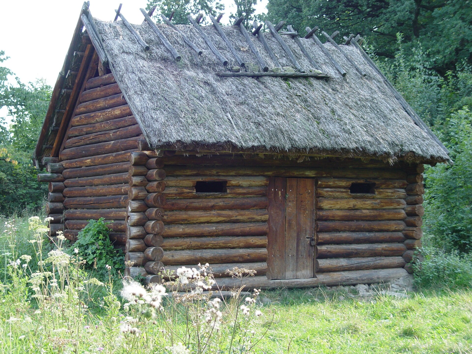 Fotografia barwna przedstawiająca chatę słowiańskich Ślężan w Rezerwacie Archeologicznym w Będkowicach. Chata jest niewielka, cała drewniana z dachem ze strzechy.