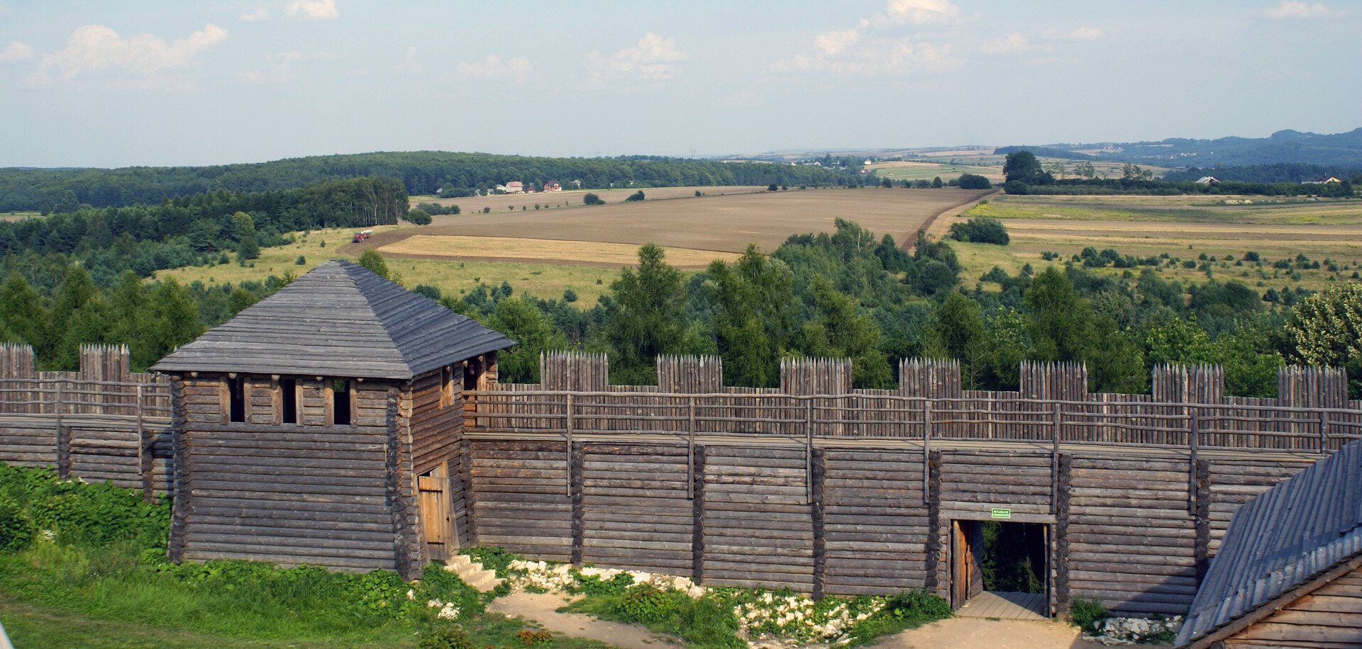 Fotografia barwna przedstawiająca Górę Birów – rekonstrukcję osady i fortyfikacji słowiańskich. W tle widać pola uprawne i lasy, na pierwszym planie stoi drewniana fortyfikacja.